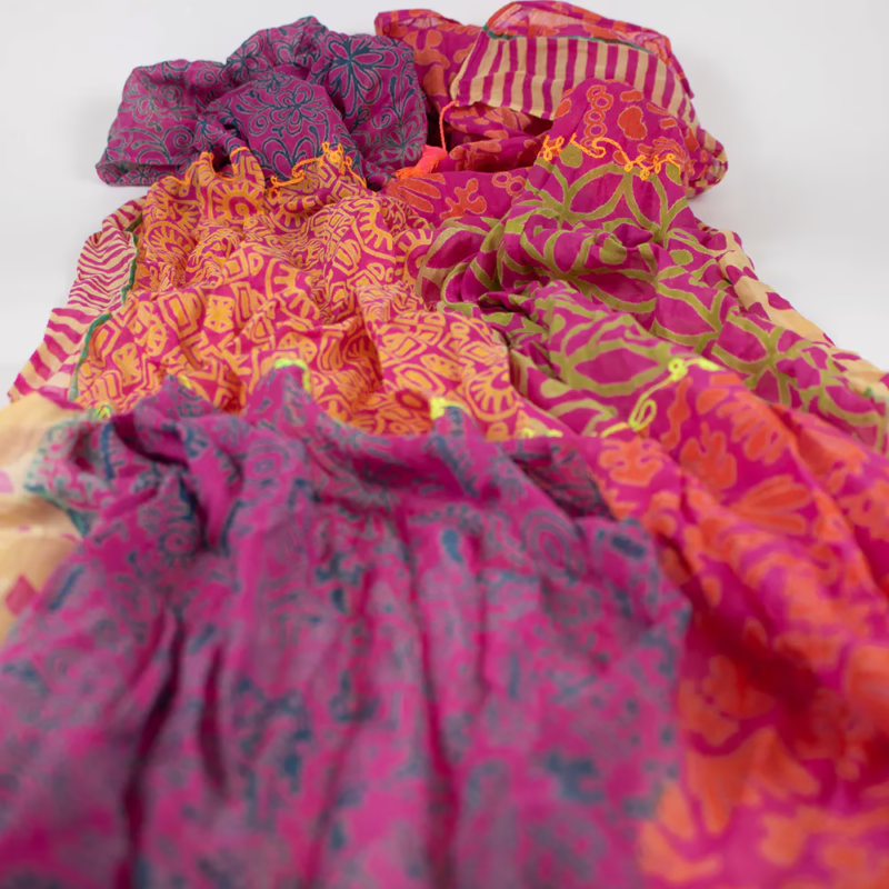 Mehrfarbiger Schal mit kombiniertem Musteraufbau. Schmale Streifen in Pink und Magenta wechseln mit Blumen- und Linienmustern in Orange und Blau. Unterschiedliche Musterflächen entlang der Länge sichtbar. Abschlüsse mit farblich abgesetzten Quasten an den Enden.